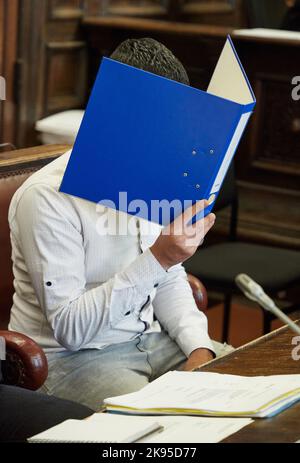 Hamburg, Germany. 26th Oct, 2022. The defendant sits in the courtroom before the trial begins, holding a blue file folder in front of his face. The 27-year-old is on trial for attempted manslaughter. The defendant is alleged to have fatally injured his partner with a knife in October 2021. He fled across adjacent rooftops and was arrested in Paris on November 11, 2021, on an international arrest warrant and transferred to Germany. Credit: Georg Wendt/dpa/Alamy Live News Stock Photo