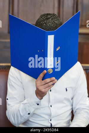 Hamburg, Germany. 26th Oct, 2022. The defendant sits in the courtroom before the trial begins, holding a blue file folder in front of his face. The 27-year-old is on trial for attempted manslaughter. The defendant is alleged to have fatally injured his partner with a knife in October 2021. He fled across adjacent rooftops and was arrested in Paris on November 11, 2021, on an international arrest warrant and transferred to Germany. Credit: Georg Wendt/dpa/Alamy Live News Stock Photo