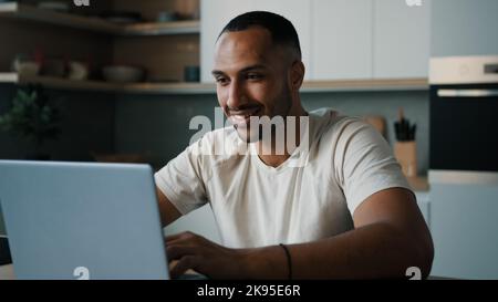 African Latina male man at kitchen at home play online game browsing in net bet working remote with laptop shopping in Internet store order delivery Stock Photo