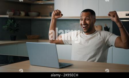 African 30s guy male man sitting at table at kitchen searching in laptop playing game online win feeling winner enjoy victory receive prize financial Stock Photo