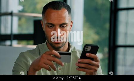 African American 30s man sitting at table indoor using smartphone for online payment Latin guy shopping buying online with mobile app and bank credit Stock Photo