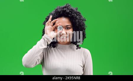 An isolated African American female on a green screen smiling and holding a silver Plated Bitcoin in front of her eye Stock Photo