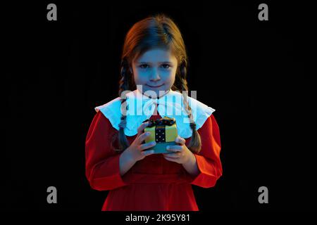 Portrait of beautiful cute little girl with pigtails wearing red festive dress standing isolated over dark background. Concept of children emotions Stock Photo