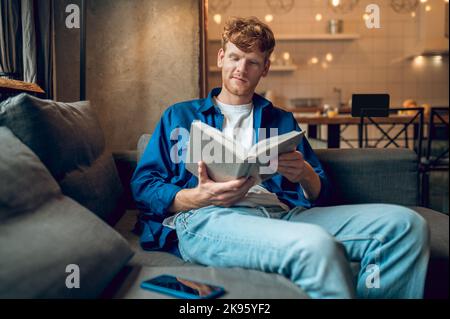 Ginger young man sitting at home at the window with a book Stock Photo