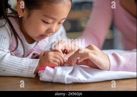 Concentrated child doing hand sewing aided by her female parent Stock Photo