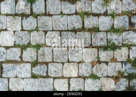 Fragment of a sidewalk paved with granite stone, with grass between the paving stones, for use as an abstract background and texture. Stock Photo