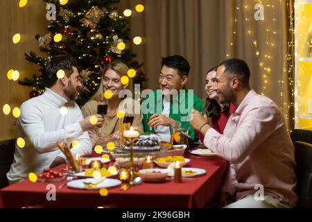 Friends celebrate Christmas eve or New Year holiday paty together sitting at the table. Feast at home group of multi ethnic festive christmas dinner. happy cheerful people clink wine glasses laugh Stock Photo