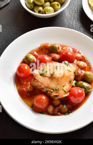 Close up view of cod stew with chickpeas, cherry tomatoes and olives on plate over dark stone background. Stock Photo
