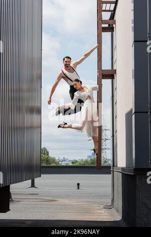 Smiling dancer looking at camera while posing near partner in skirt and heels on ladder on roof Stock Photo