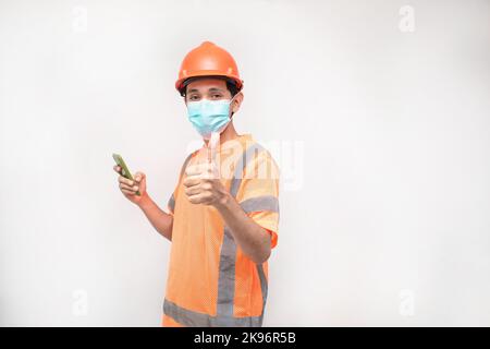 A Caucasian male wearing a construction helmet and giving a thumbs-up while using his smartphone Stock Photo