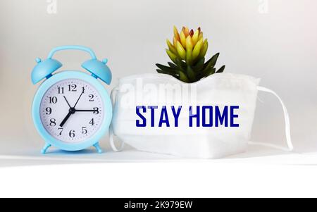 Stay at home save lives phrases on a medical mask on a white background with a clock and a house flower. Take care of yourself, stay at home. Stock Photo