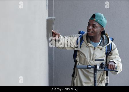 Side view of food delivery boy ringing doorbell while riding electric scooter Stock Photo