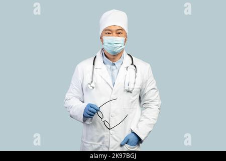 Confident mature korean man doctor in white coat, protective gloves, mask takes off glasses Stock Photo