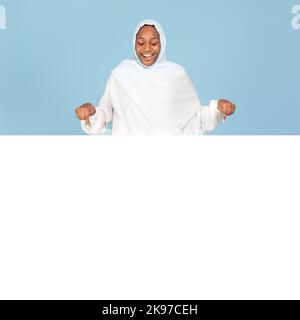 Excited black woman in hijab pointing down at white empty board for advertisement over blue background, free space Stock Photo