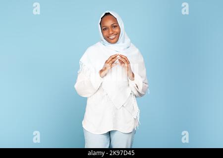 Portrait of black muslim woman joining fingers together while posing over blue studio background and smiling to camera Stock Photo