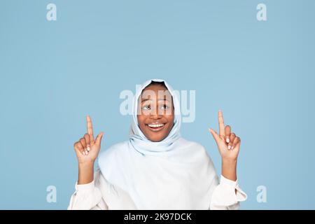 Happy african american muslim woman pointing with both hands and looking up at copy space, showing space for your ad Stock Photo