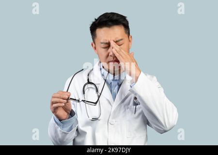 Tired sad middle aged chinese man doctor in white coat takes off glasses, rubs eyes with hand Stock Photo