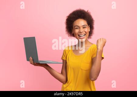 Cheerful excited young mixed race woman with curly hair rejoices online success with laptop, making victory gesture Stock Photo