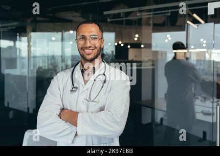 Portrait of a young handsome male hispanic doctor. Standing in the glass hall of the hospital wearing glasses, a white coat and a stethoscope. He looks at the camera, crosses his arms, smiles. Stock Photo