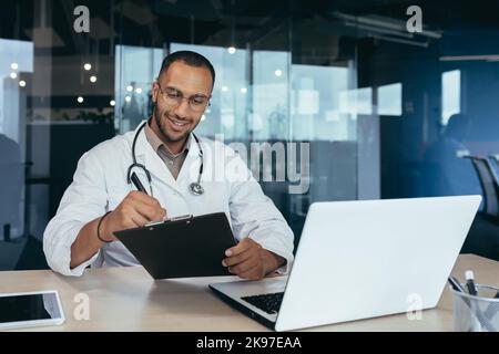 Online reception at the hospital. Young handsome hispanic doctor talking to patient via laptop. He sits in the office at the work table, writes down the history of the cough, the patient's complaints, and the prescription for medicine. Stock Photo