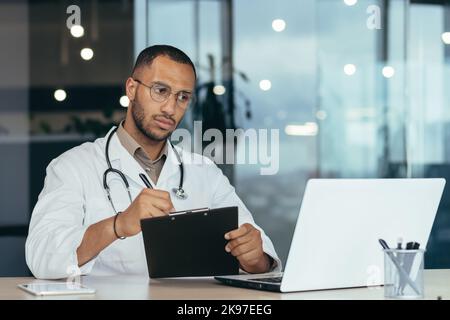 A young handsome man, a Latin American student, a trainee doctor, an intern studies via video link from a laptop. He sits serious in the office, passes the exam, writes down. Stock Photo