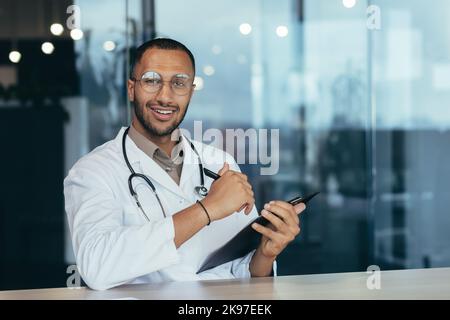 Portrait of a young handsome Latin American doctor. Sitting in the office, holding a pen with documents in his hands, looking at the camera, smiling Stock Photo