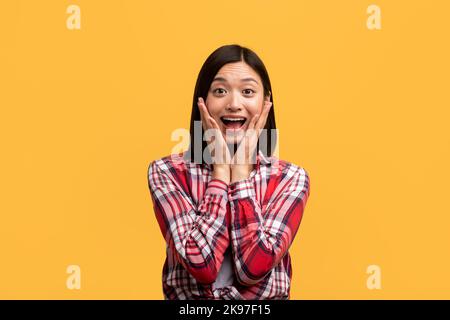 Unbelievable. Shocked asian lady with open mouth and hands on cheeks, emotionally reacting to news, yellow background Stock Photo