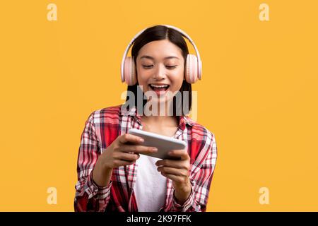 Entertainment and online games. Joyful asian lady in modern wireless headphones playing on smartphone Stock Photo