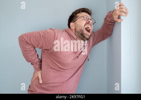 European man is suffering from severe back pain, he is screaming while holding hand against the wall Stock Photo