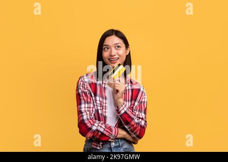 Shopping time concept. Dreamy asian lady holding credit card and looking away, thinking what to buy, copy space Stock Photo