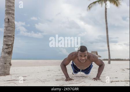 Muscular African American athlete in sportswear pushing up while training on sandy beach under cloudy sky and looking at camera Stock Photo