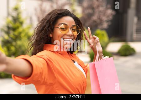 Glad millennial black woman in casual and glasses with bags takes selfie on smartphone, make peace sign with hand Stock Photo