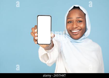 Excited black woman in hijab demonstrating smartphone with white blank screen, posing over blue background, mockup Stock Photo