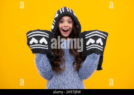 Warm hat with hood and scarf. Amazed teenager. Modern teenage girl 12, 13, 14 year old wearing sweater and knitted hat on isolated yellow background Stock Photo