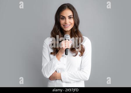 Beautiful business woman is speaking on conference. Stylish girl singing songs with microphone, holding mic at karaoke, posing against gray background Stock Photo