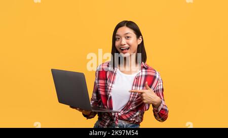 Happy chinese woman using laptop and pointing finger at pc computer, working online and smiling at camera, panorama Stock Photo
