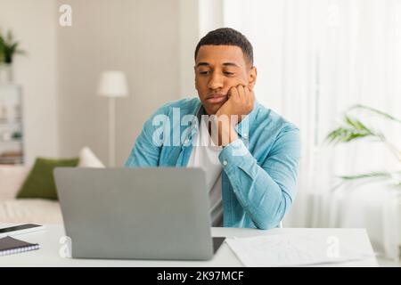 Sad tired bored young black guy in casual looks at laptop in light living room office interior, empty space Stock Photo
