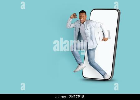 Cheerful excited millennial black man with open mouth with laptop jumping out of huge smartphone Stock Photo