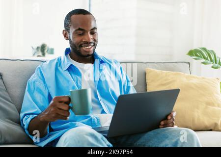 Portrait of smiling black man using laptop, holding cup and drinking morning coffee, sitting on sofa and reading news Stock Photo