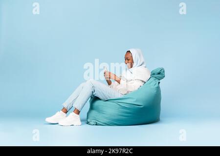 Positive black muslim woman chilling in beanbag chair with smartphone, chatting with friends in social networks Stock Photo