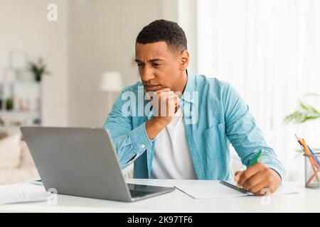 Serious concentrated young black man in jeans looking at laptop, making notes, watching online lesson Stock Photo