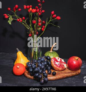 Still life with fruits and rosehip branches in a glass vase on black background. Thanksgiving concept. Closeup. Stock Photo