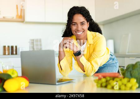 Searching for new healthy recipes. Happy african american woman cooking fresh salad in modern kitchen, using laptop Stock Photo