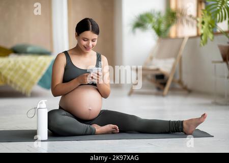 Pregnant woman relaxing with smartphone on yoga mat after training at home Stock Photo