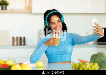 Happy black woman in sport clothes taking selfie with fresh juice, sitting at table in kitchen interior, free space Stock Photo