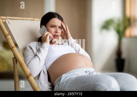 Worried Pregnant Woman Talking On Cellphone While Sitting In Chair At Home Stock Photo