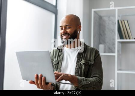 Happy male freelancer using laptop computer and wearing earbuds, standing near window and typing on keyboard Stock Photo