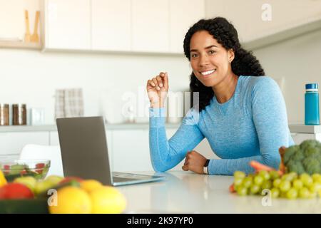 Happy fit black lady using laptop browsing internet, searching recipes for healthy dinner, sitting in kitchen at home Stock Photo