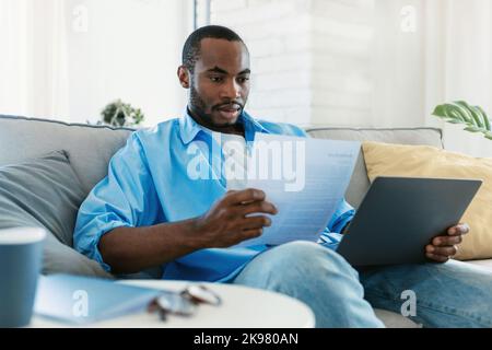 Paperwork concept. Portrait of black man working at home, reading paper or financial document, sitting on sofa Stock Photo