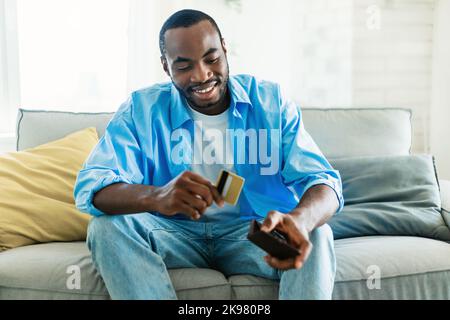 Excited african american man selecting credit card from his card wallet, sitting on sofa in living room interior Stock Photo